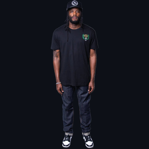 A man wearing a black T-shirt with embroidery details on the front, paired with blue jeans and a cap.